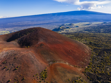 Aerial view of Mauna Kea and a reddish volcanic cinder cone surrounded by sparse vegetation and lava fields under a blue sky in Hawaii County Hawaii United States.
