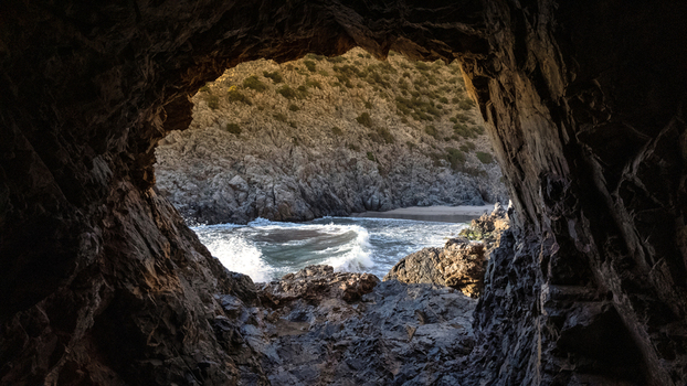 View of Spiaggia di Cala Domestica through a dark sea cave opening with turquoise waves crashing against rugged cliffs Provincia del Sulcis Iglesiente Sardegna Italy. View of Spiaggia di Cala Domestica through a dark sea cave opening with turquoise waves crashing against rugged cliffs Provincia del Sulcis Iglesiente Sardegna Italy.