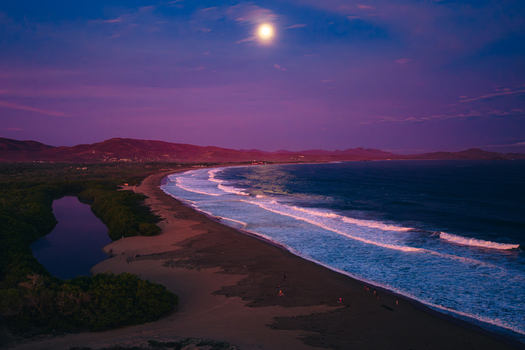 Aerial view of Playa Punta Perula under a bright full moon with gentle waves washing onto the dark sandy beach and lush coastal vegetation in Perula Jalisco Mexico. Aerial view of Playa Punta Perula under a bright full moon with gentle waves washing onto the dark sandy beach and lush coastal vegetation in Perula Jalisco Mexico.