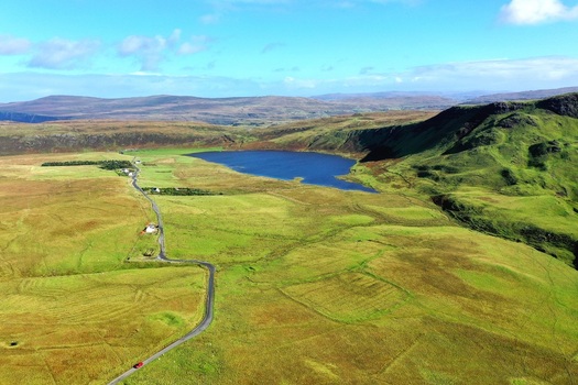 Aerial view of a winding road leading through lush green hills toward a dark blue lake in Isle of Skye Scotland United Kingdom.