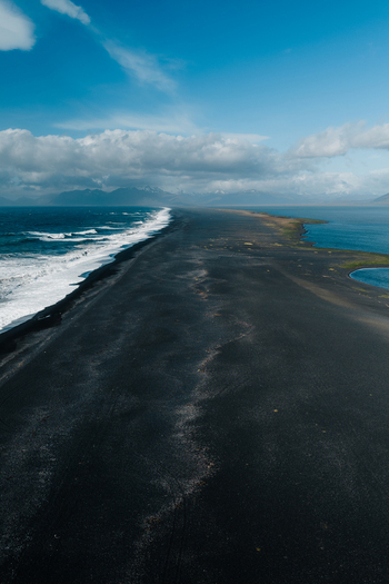 Aerial view of Stokksnes black sand beach with crashing waves dark volcanic sand and distant mountains under a cloudy blue sky in Iceland.