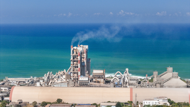 Aerial view of a large industrial cement factory complex with smoking chimneys and storage silos situated along the coastline of Indonesia.