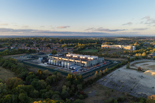 Aerial view of a modern data center facility surrounded by lush greenery and trees under a soft golden hour sky in the United Kingdom. Aerial view of a modern data center facility surrounded by lush greenery and trees under a soft golden hour sky in the United Kingdom.