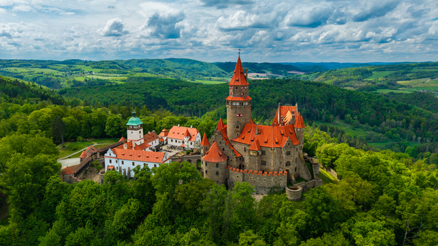 Aerial view of Bouzov Castle with its red tiled roofs and tall towers nestled within a dense green forest under a cloudy sky in Bouzov Olomoucky kraj Czechia. Aerial view of Bouzov Castle with its red tiled roofs and tall towers nestled within a dense green forest under a cloudy sky in Bouzov Olomoucky kraj Czechia.