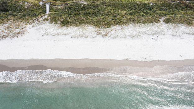 Aerial view of the white sand beach and turquoise water at Omaha Beach with a wooden walkway leading through the dunes in Omaha Auckland New Zealand.