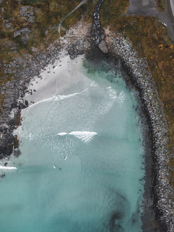 Aerial view of surfers in the turquoise waters of a beach in Lofoten surrounded by rocky cliffs and rugged coastline in Svolvaer Nordland Norway.