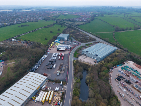 Aerial view of industrial buildings and vehicles nestled amongst lush green fields and tree lines with a small pond reflecting the overcast sky Redditch United Kingdom. Aerial view of industrial buildings and vehicles nestled amongst lush green fields and tree lines with a small pond reflecting the overcast sky Redditch United Kingdom.