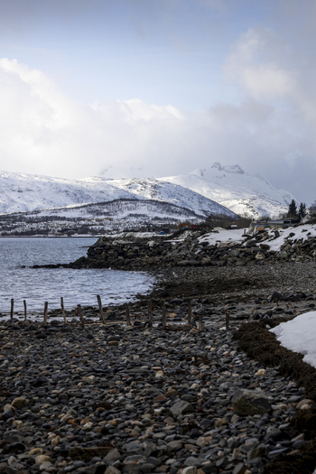 View of the rocky shoreline and snow covered mountains under a cloudy sky Kvaloya Troms Norway. View of the rocky shoreline and snow covered mountains under a cloudy sky Kvaloya Troms Norway.