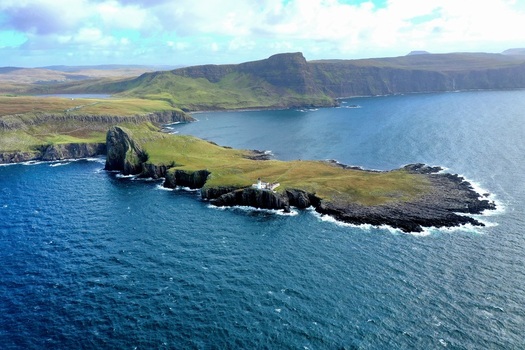Aerial view of Neist Point Lighthouse on a dramatic rocky cliff edge surrounded by deep blue ocean waters featuring rugged coastal terrain Isle of Skye Scotland United Kingdom.