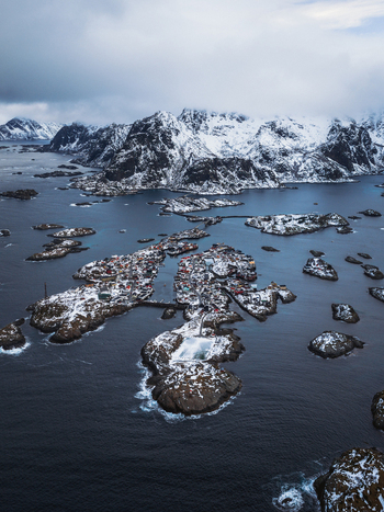 Aerial view of a snow dusted archipelago where the quaint village nestles amidst the rugged icy landscape and the dark swirling sea Henningsv&aelig;r Nordland Norway.
