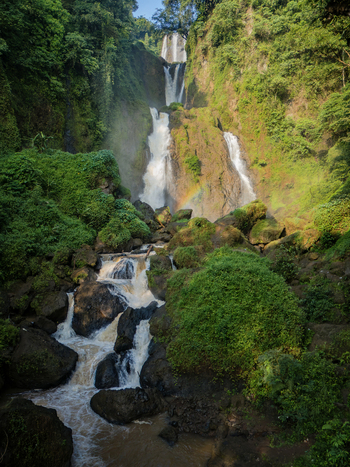 Aerial view of Curug Telu waterfall with multi tiered cascades lush tropical forest and a faint rainbow in the mist in Kendal Central Java Indonesia. Aerial view of Curug Telu waterfall with multi tiered cascades lush tropical forest and a faint rainbow in the mist in Kendal Central Java Indonesia.