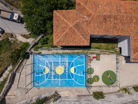 Aerial view of a blue basketball court and a small playground next to a building with terracotta tiled roofs and lush green trees in Dhermi Vlore County Albania.