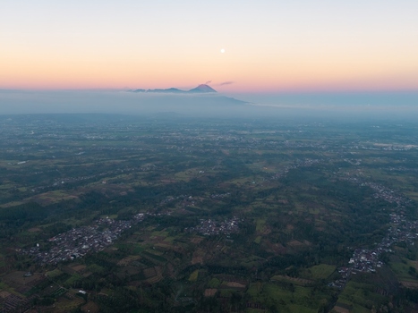 Aerial view of the landscape near Malang with a distant volcano rising above clouds during sunset featuring scattered settlements and agricultural fields in Malang East Java Indonesia. Aerial view of the landscape near Malang with a distant volcano rising above clouds during sunset featuring scattered settlements and agricultural fields in Malang East Java Indonesia.