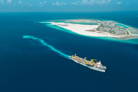 Aerial view of a large industrial dredger ship sailing through deep blue ocean waters towards a tropical island with white beaches in Vashafaru Haa Alif Atoll Maldives. Aerial view of a large industrial dredger ship sailing through deep blue ocean waters towards a tropical island with white beaches in Vashafaru Haa Alif Atoll Maldives.