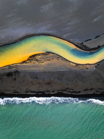 Aerial view of a winding glacial river with vibrant yellow and blue waters flowing across dark volcanic sand into the ocean in Iceland. Aerial view of a winding glacial river with vibrant yellow and blue waters flowing across dark volcanic sand into the ocean in Iceland.