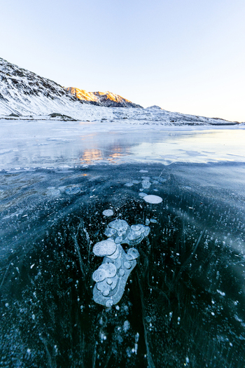 View of methane bubbles trapped in the frozen surface of a lake with snow covered mountains under a clear sky in Madesimo Lombardy Italy. View of methane bubbles trapped in the frozen surface of a lake with snow covered mountains under a clear sky in Madesimo Lombardy Italy.