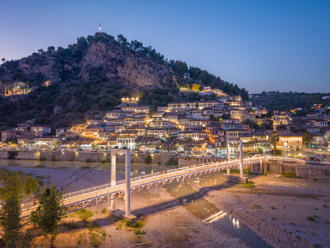 Aerial view of the historic town of Berat with traditional Ottoman style houses illuminated at dusk featuring the Osum River and a pedestrian bridge in Berat Berat County Albania.