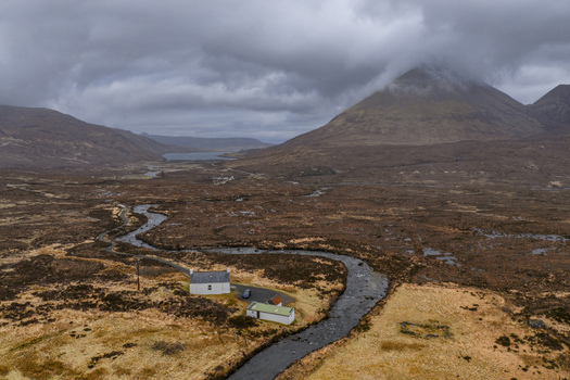 Aerial view of a winding river flowing through the rugged moorland near a small white cottage under a dramatic cloudy sky in Isle of Skye Scotland United Kingdom. Aerial view of a winding river flowing through the rugged moorland near a small white cottage under a dramatic cloudy sky in Isle of Skye Scotland United Kingdom.