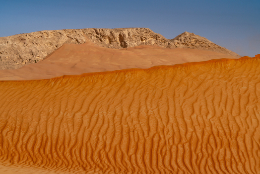 View of red sand dunes with wind ripples and a rocky mountain ridge under a clear blue sky in the desert Dubai Dubai United Arab Emirates. View of red sand dunes with wind ripples and a rocky mountain ridge under a clear blue sky in the desert Dubai Dubai United Arab Emirates.