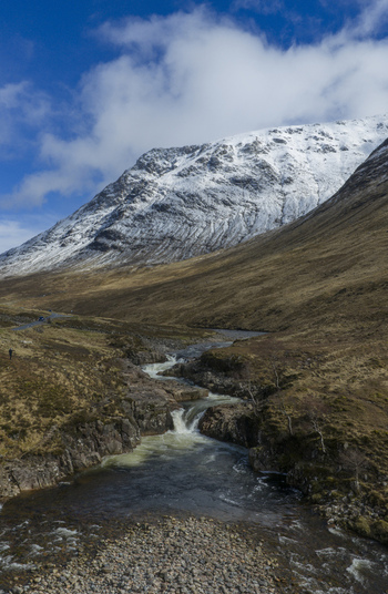 Aerial view of Glen Etive Waterfall flowing through a rugged valley with snow capped mountains under a blue sky Ballachulish Scotland United Kingdom.