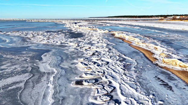 Aerial view of a frozen coastline with ice floes scattered across the water and a snow covered sandy beach under a clear blue sky in Latvia. Aerial view of a frozen coastline with ice floes scattered across the water and a snow covered sandy beach under a clear blue sky in Latvia.