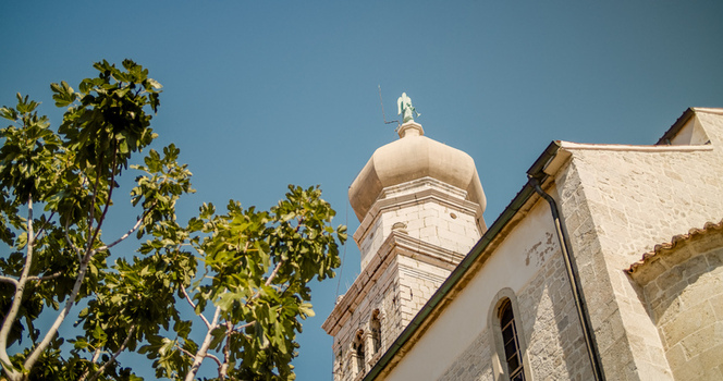 Krk Primorje Gorski Kotar Croatia 19 September 2026 View of the stone bell tower of the Cathedral of the Assumption with a dome and angel statue framed by fig tree branches. Krk Primorje Gorski Kotar Croatia 19 September 2026 View of the stone bell tower of the Cathedral of the Assumption with a dome and angel statue framed by fig tree branches.