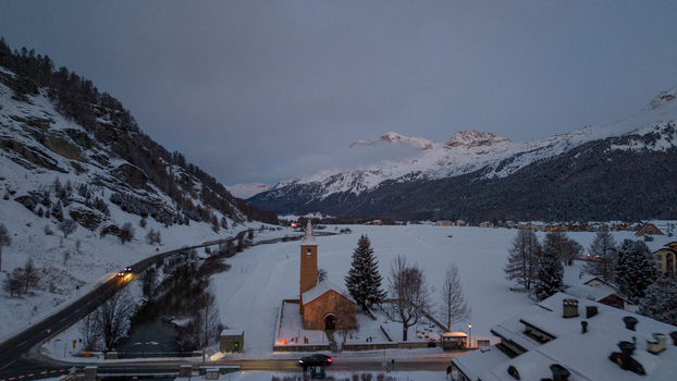 Aerial view of the historic church in Sils Maria surrounded by snow covered mountains and a winding road at dusk in Sils im EngadinSegl Grisons Switzerland.