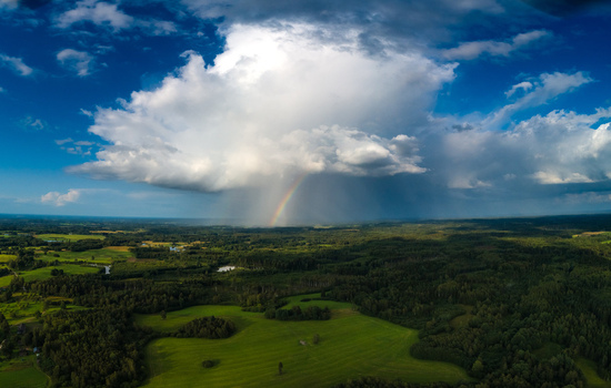 Aerial view of a vibrant rainbow arching over a lush green landscape with dense forests rolling fields and a dramatic storm cloud in Latvia. Aerial view of a vibrant rainbow arching over a lush green landscape with dense forests rolling fields and a dramatic storm cloud in Latvia.