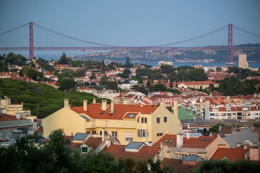 Aerial view of the 25 April Bridge spanning the Tagus River with a dense residential neighborhood featuring red tiled roofs and lush trees in Lisboa Lisboa Portugal.