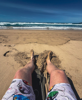 View of a person s legs and sandy feet resting on the shore with ocean waves and a clear blue sky at Plettenberg Bay Western Cape South Africa. View of a person s legs and sandy feet resting on the shore with ocean waves and a clear blue sky at Plettenberg Bay Western Cape South Africa.