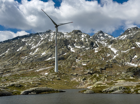 View of a wind turbine in the rugged alpine landscape of Sankt Gotthard Pass with snow capped peaks and a small mountain lake under a cloudy sky Airolo Ticino Switzerland.