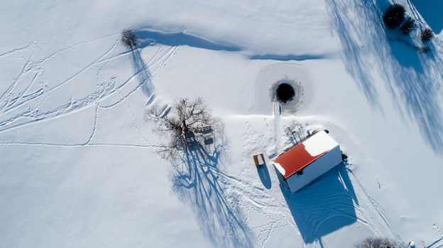 Aerial view of a red roofed building situated in a vast snow covered landscape with a large tree casting long shadows and a circular hole in the ice in Latvia. Aerial view of a red roofed building situated in a vast snow covered landscape with a large tree casting long shadows and a circular hole in the ice in Latvia.