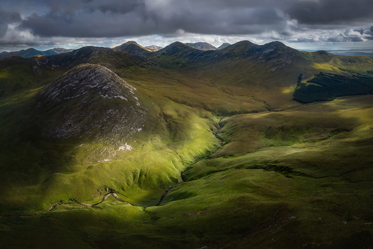 Aerial view of Diamond Hill and the Twelve Bens mountain range under dramatic storm clouds with sunlight hitting the green valley County Galway Ireland. Aerial view of Diamond Hill and the Twelve Bens mountain range under dramatic storm clouds with sunlight hitting the green valley County Galway Ireland.