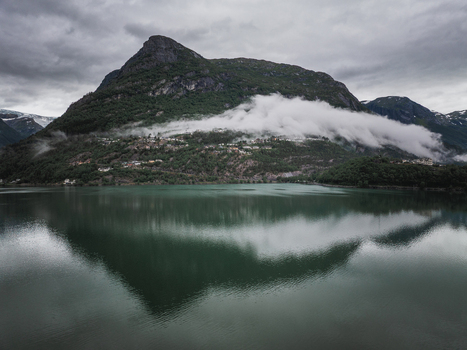 Aerial view of a mountain reflecting in a calm lake with low hanging clouds and lush green slopes in Otta Innlandet Norway.