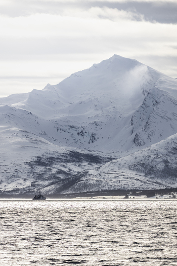 View of snow capped mountains rising above a calm fjord with a small boat sailing across the water in Tromso Troms Norway.