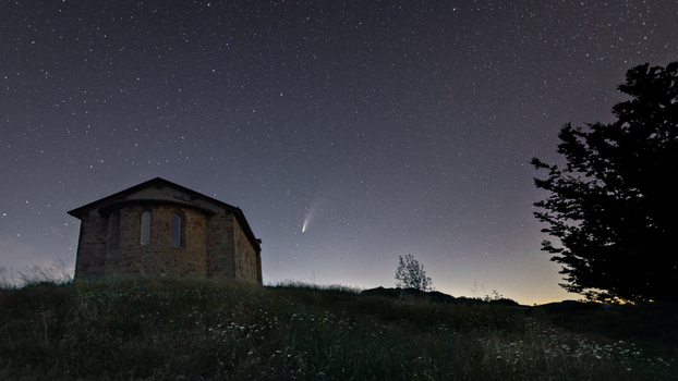 View of a small stone chapel on a grassy hill under a starry night sky featuring a bright comet with a visible tail and silhouetted trees. View of a small stone chapel on a grassy hill under a starry night sky featuring a bright comet with a visible tail and silhouetted trees.