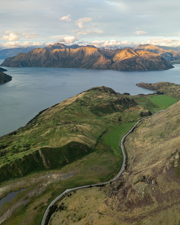 Aerial view of the winding road snaking through the lush green hills towards the serene lake surrounded by majestic mountains Glendhu Bay Otago Region New Zealand. Aerial view of the winding road snaking through the lush green hills towards the serene lake surrounded by majestic mountains Glendhu Bay Otago Region New Zealand.