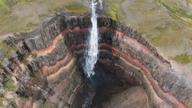 Aerial view of Hengifoss waterfall cascading down layered cliffs with striking red clay bands highlighting the dark basalt rock Iceland.