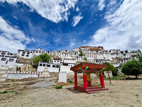 View of Thiksey Monastery with its white buildings terraced on the hillside and a vibrant red prayer wheel structure in the foreground under a blue sky in Thiksey Ladakh India.