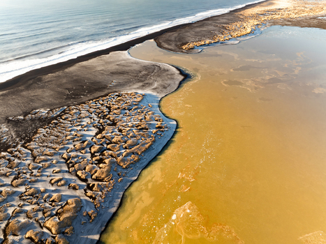 Aerial view of the rugged coastline in Iceland featuring black sand beaches textured terrain and calm yellowish coastal waters in Reykjanesbaer Iceland. Aerial view of the rugged coastline in Iceland featuring black sand beaches textured terrain and calm yellowish coastal waters in Reykjanesbaer Iceland.