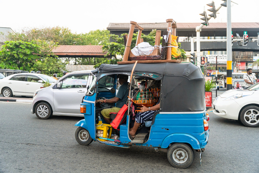 Sri Lanka 15 January 2026 View of a blue tuk tuk transporting a large wooden table on its roof through city traffic with cars and trees in the background.