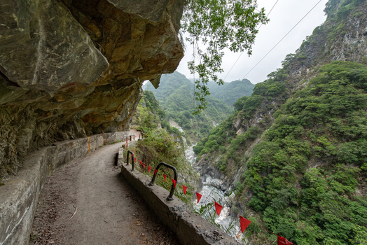 View of the narrow hiking trail carved into the cliffside of Taroko National Park with lush green mountains and a river gorge below in Xiulin Township Hualien County Taiwan.