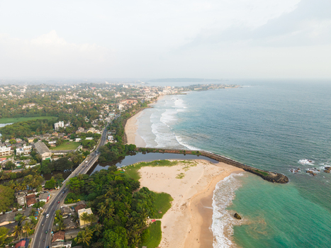 Aerial view of the coastline featuring a sandy beach coastal road and lush tropical greenery under a soft hazy sky in Galle Southern Province Sri Lanka.
