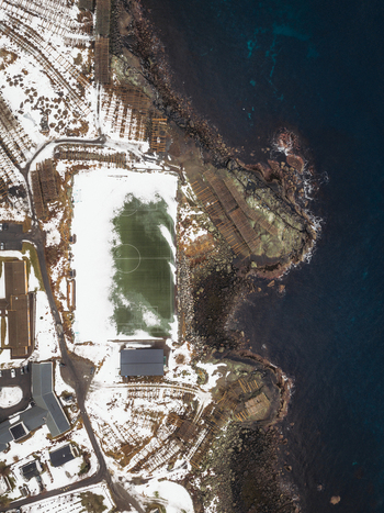 Aerial view of a snow dusted landscape meeting the dark waters where a soccer field stands as a bright green island Reinebringen Nordland Norway.