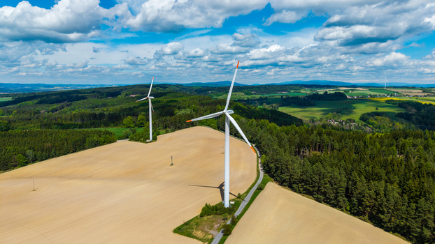Aerial view of wind turbines standing on a golden field surrounded by lush green forests under a cloudy blue sky Gruna Pardubicky kraj Czechia. Aerial view of wind turbines standing on a golden field surrounded by lush green forests under a cloudy blue sky Gruna Pardubicky kraj Czechia.