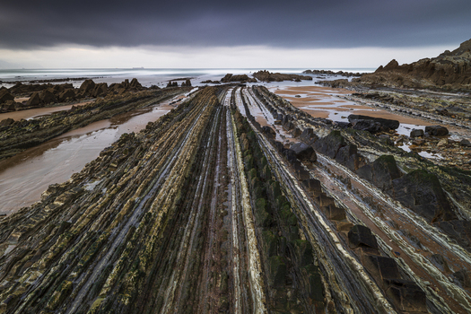 View of Playa de Sopelana featuring unique flysch rock formations under a dramatic cloudy sky in Vizcaya Pais Vasco Spain. View of Playa de Sopelana featuring unique flysch rock formations under a dramatic cloudy sky in Vizcaya Pais Vasco Spain.