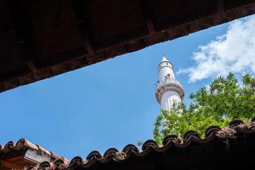 View of the white minaret of a mosque framed by traditional wooden eaves and terracotta roof tiles under a blue sky Kruje Durres County Albania.