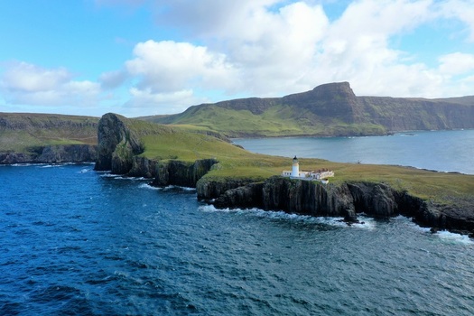 Aerial view of Neist Point Lighthouse perched on rugged cliffs above the deep blue sea with grassy headlands under a cloudy sky in Isle of Skye Scotland United Kingdom.