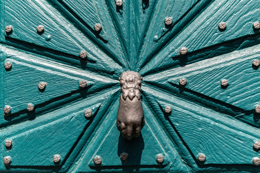 View of an ornate teal wooden door featuring a starburst pattern with metal studs and a hand shaped iron door knocker in the historic village of Monreal Rhineland Palatinate Germany. View of an ornate teal wooden door featuring a starburst pattern with metal studs and a hand shaped iron door knocker in the historic village of Monreal Rhineland Palatinate Germany.
