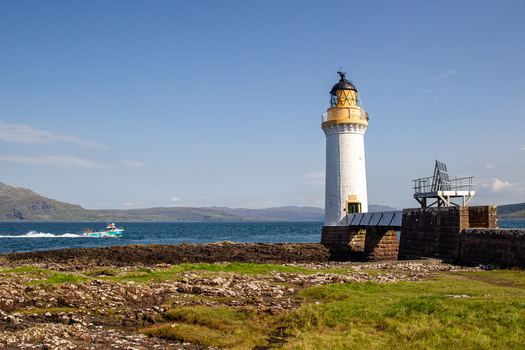 View of Tobermory Lighthouse standing on a rocky coastline with a boat passing by on the blue sea under a clear sky Isle of Mull Scotland United Kingdom. View of Tobermory Lighthouse standing on a rocky coastline with a boat passing by on the blue sea under a clear sky Isle of Mull Scotland United Kingdom.